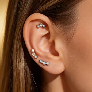 May include: Close-up of an ear adorned with multiple pieces of jewelry. Three small, curved earrings are visible, each featuring a series of sparkling, clear gemstones. The earrings are set in a light-colored metal, creating a delicate and elegant look.