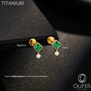 Puede incluir: Un par de pendientes de botón dorados. Cada pendiente presenta una gema cuadrada de color verde esmeralda, una pequeña gema blanca y un poste. Los pendientes miden 8 mm de largo. La palabra "TITANIUM" está en la parte superior.