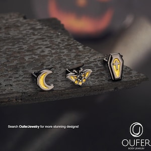 May include: Three pieces of body jewelry with a Halloween theme. The pieces include a crescent moon, a moth, and a coffin, all with black and gold accents. The image also includes the text "Search OuferJewelry for more stunning designs!"