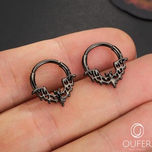 Puede incluir: Dos pendientes de aro negros con un diseño de ala de murciélago. Los pendientes son de metal y tienen un cierre de bisagra. Los pendientes se muestran en una mano.