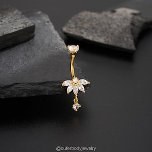 May include: A gold-colored belly button ring with a floral design. The ring features a curved barbell with a clear gem at the top and a flower-shaped cluster of clear gems at the bottom. A small, dangling gem hangs below the flower.