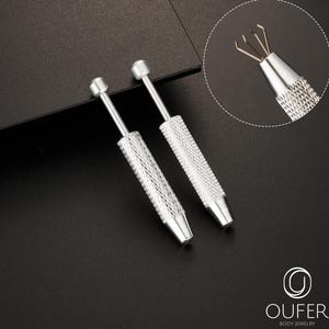 Puede incluir: Dos herramientas plateadas para joyería corporal con mangos texturizados y una punta en forma de garra. Las herramientas están sobre una superficie oscura, con una incrustación de primer plano que muestra la punta de la garra. De Oufer Body Jewelry.