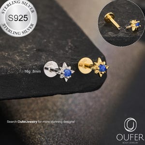 Könnte beinhalten: Zwei sternförmige Körperschmuck-Ohrstecker. Einer ist silberfarben mit einem blauen Edelstein, der andere ist goldfarben mit einem blauen Edelstein. Das Bild enthält eine Nahaufnahme des Schmucks und ein Logo mit der Aufschrift "OUFER BODY JEWELRY". Der Text lautet "STERLING SILVER S925".