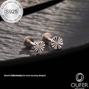 May include: Two silver body jewelry studs. One stud is heart-shaped with radiating lines, and the other is a circular design with similar lines. The studs are on a dark, textured surface. The image includes the text "STERLING SILVER S925".