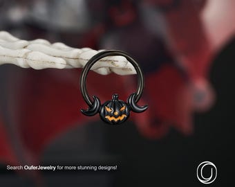 Puede incluir: Anillo de joyería corporal negro con temática de Halloween con un diseño de calabaza tallada con acentos de media luna. La calabaza tiene un brillo naranja. El anillo está sostenido por una mano esquelética blanca. El texto "Search OuferJewelry for more stunning designs!" es visible.