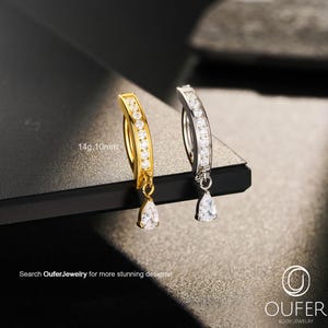 Könnte beinhalten: Zwei gebogene Bauchnabelpiercings, eines goldfarben und eines silberfarben, jeweils mit einer Reihe klarer Steine in Kanalfassung entlang des Bogens und einem tropfenförmigen Anhänger. Der Text 14g 10mm ist beim goldenen Ring sichtbar.