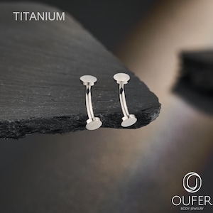 Op de afbeelding: Twee zilverkleurige, gebogen titanium piercingsieraden met platte, ronde uiteinden. De tekst TITANIUM is zichtbaar in de linkerbovenhoek en de tekst OUFER BODY JEWELRY is zichtbaar in de rechterbenedenhoek van de afbeelding.
