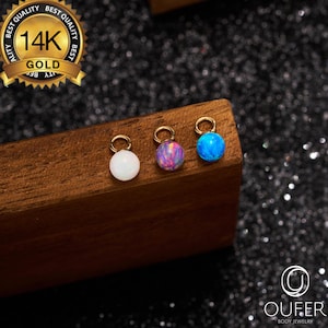Puede incluir: Tres pequeños colgantes redondos de joyería corporal en colores ópalo blanco, morado-rosa y azul. Cada colgante tiene un lazo dorado en la parte superior. Los colgantes se exhiben sobre un bloque de madera, con un sello dorado que dice "14K GOLD".