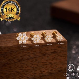 May include: Four gold flower-shaped earrings with clear gemstones, ranging in size from 3.3mm to 6.5mm. The earrings are displayed on a wooden block, with a gold seal that reads "14K GOLD". The brand name "OUFER BODY JEWELRY" is also visible.