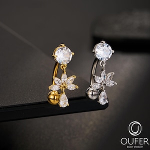May include: Two belly button rings, one gold-toned and one silver-toned, each featuring a large, clear, round gemstone at the top. Below, a floral design with clear gemstones and a teardrop-shaped gem hangs from the curved bar. A small, round gem is at the bottom.