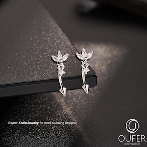 Può includere: Orecchini in argento con design floreale e pendenti a forma di freccia. Gli orecchini presentano pietre preziose trasparenti e sono esposti su una superficie scura. L'immagine include il testo "Search OuferJewelry for more stunning designs!"