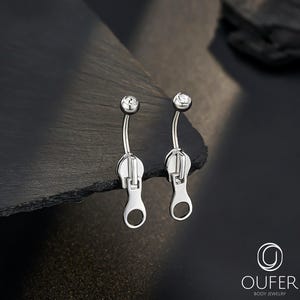 Puede incluir: Un par de piercings para el ombligo de color plateado. Cada uno presenta una barra curva con una pequeña gema redonda y transparente en la parte superior y un dije de cremallera en la parte inferior. El fondo es oscuro con una superficie texturizada.