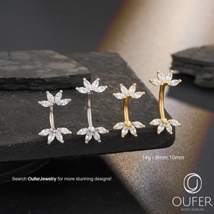 Pode incluir: Três piercings para o umbigo com um design floral. Dois são prateados e um dourado. Cada anel apresenta uma barra curva com um grupo de pedras transparentes com corte marquesa em cada extremidade. O texto "Search OuferJewelry for more stunning designs!" é visível.