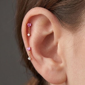 May include: Two gold cartilage earrings featuring a small pink heart-shaped gemstone attached to a delicate chain that ends in a single clear round gemstone. These earrings are positioned vertically along the upper edge of the ear cartilage.