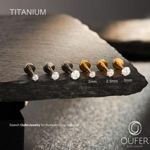 May include: A set of six titanium labret studs with clear round gemstones. Three studs are silver-toned and three are gold-toned, with sizes labeled as 2mm, 2.5mm, and 3mm. The text TITANIUM and OUFER BODY JEWELRY are visible on the image.