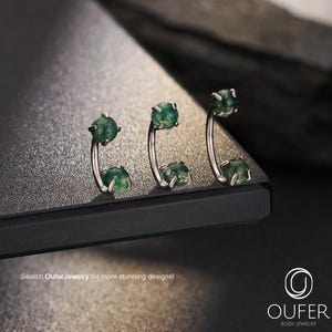 May include: Three curved barbell body jewellery pieces, each featuring two prong-set round green moss agate stones. The metal is silver-toned. The text "Search OuferJewelry for more stunning designs!" and the "OUFER BODY JEWELRY" logo are visible.