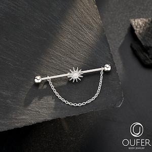 May include: A silver industrial barbell body piercing. The central design is a sunburst, with a dangling chain. The barbell has a ball on each end. The jewellery is displayed on a dark, textured surface.