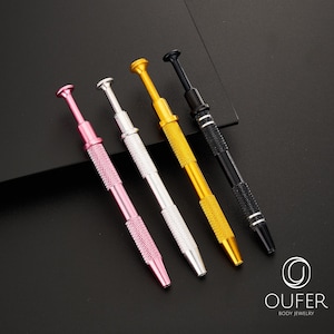 May include: Four colorful body jewelry tools in pink, silver, gold, and black. Each tool has a textured handle and a small, rounded tip. The tools are arranged diagonally against a dark background, with the brand name "OUFER BODY JEWELRY" visible.