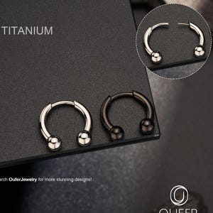 May include: Two circular titanium horseshoe rings with ball ends, one in silver tone and one in black. The text TITANIUM is visible in the upper left corner, and the Oufer Jewelry logo and text are in the bottom right corner of the image.