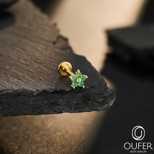 Puede incluir: Un pendiente tipo labret de oro con un diseño de flor en la parte superior, adornado con cinco pequeñas piedras preciosas redondas de color verde. La pieza tiene un poste plano y el logotipo OUFER BODY JEWELRY es visible.