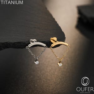 May include: Two titanium body jewellery pieces, one silver-toned and one gold-toned, each featuring a curved bar, a delicate chain, and a small clear gemstone charm. The text TITANIUM and OUFER BODY JEWELRY are visible in the image.