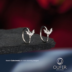 May include: A pair of silver-toned hoop earrings, each featuring a small, flat bat-shaped charm attached to the circular band. The text "Search OuferJewelry for more stunning designs!" and the "OUFER BODY JEWELRY" logo are visible at the bottom.