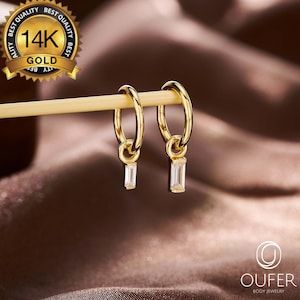 May include: A pair of gold hoop earrings with rectangular, clear gemstones hanging from the hoops. The earrings are displayed on a light-colored stick against a blurred, brown background. A gold seal reads "14K GOLD".