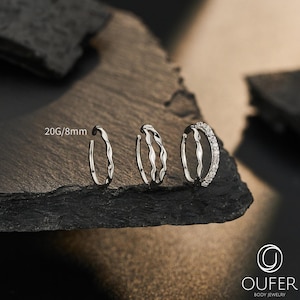 Puede incluir: Tres aros plateados con la etiqueta 20G/8mm. El primer anillo es un aro fino simple, el segundo presenta un diseño ondulado y el tercero es un aro ondulado adornado con una hilera de pequeñas piedras transparentes. La marca OUFER es visible.