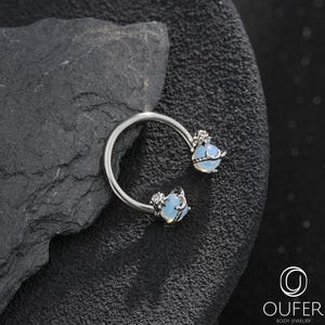 Puede incluir: Una joya corporal en forma de herradura de tono plateado con dos piedras redondas de color azul claro iridiscente en cada extremo. Cada piedra está acentuada por una pequeña y delicada luna creciente y una diminuta gema transparente. El texto OUFER BODY JEWELRY es visible.