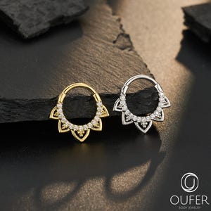 Peut inclure: Deux anneaux segmentaires circulaires, l'un doré et l'autre argenté, chacun doté d'une attache décorative en forme d'éventail ornée de petites pierres claires. Les bijoux sont présentés ensemble et le logo OUFER BODY JEWELRY est visible.