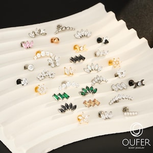 May include: A collection of body jewelry, including earrings and studs, displayed on a white, textured surface. The jewelry features various designs with clear, black, green, and pink gemstones, as well as gold and silver tones. The brand name "OUFER BODY JEWELRY" is visible.