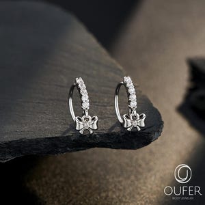 Peut inclure: Une paire de boucles d'oreilles créoles argentées avec un motif de nœud. Chaque boucle d'oreille présente une rangée de petits pierres précieuses transparentes le long de l'anneau et un pendentif en forme de nœud avec une seule pierre précieuse. Le logo 'OUFER' est visible.