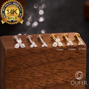 May include: A collection of 14K gold body jewelry, including lip and cartilage studs. Each stud features a cluster of clear, sparkling gemstones. Sizes are 4.8mm, 3.3mm, and 2.7mm. The jewelry is displayed on a wooden block. The logo for OUFER Body Jewelry is visible.