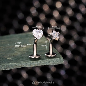 Könnte beinhalten: Zwei Schmuckstücke für den Körper. Eines ist ein herzförmiger Stecker, 4,5 mm breit, und das andere ist ein sternförmiger Stecker, 4,6 mm breit. Beide sind silberfarben mit klaren Edelsteinen. Die Stifte sind 8 mm lang und 1,6 mm dick.