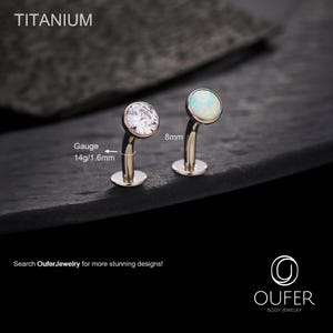 Puede incluir: Dos piezas de joyería corporal de titanio. Una presenta una gema clara y facetada, la otra un ópalo. El calibre es de 1,6 mm y la longitud es de 8 mm. La palabra "TITANIUM" está en la parte superior. El texto "Search OuferJewelry for more stunning designs!" está en la parte inferior.