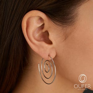 May include: Silver-toned spiral hoop earrings. The earrings feature a three-tiered spiral design, with each tier decreasing in size towards the center. The earrings are worn on an ear, with the ear and part of the head visible in the background. The earrings are a simple, elegant design.