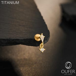 May include: A gold-tone titanium flat back stud earring featuring a kite-shaped clear gemstone at the top and a small dangling round clear gemstone charm. The text TITANIUM and OUFER BODY JEWELRY are visible in the image.