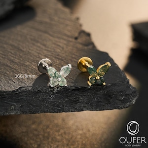 Puede incluir: Dos piercings labret con forma de mariposa, uno en metal plateado y otro en dorado, con alas de piedra ágata musgosa verde. El texto 16G/8mm es visible cerca del piercing plateado y la marca OUFER BODY JEWELRY aparece impresa debajo.