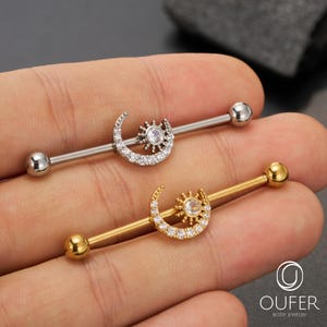 Pode incluir: Dois piercings industriais com desenhos celestiais. Um é prateado, com um design de lua crescente e sol com pedras transparentes. O outro é dourado, com um design semelhante. Ambos têm uma bola em cada extremidade.