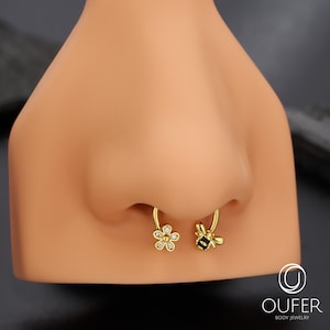 May include: Gold septum jewellery with a flower and bee design. The flower has clear gem accents, and the bee has black and yellow details. The jewellery is displayed on a mannequin nose.