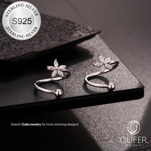 Peut inclure: Deux ear cuffs argentés avec un motif floral et des détails scintillants. Les ear cuffs ont une forme en spirale avec une petite boule à l'extrémité. L'image comprend également un logo "Oufer Jewelry".