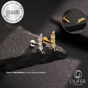 May include: Two feather-shaped body jewelry studs, one silver and one gold, each adorned with small, sparkling stones. The silver stud is on the left, and the gold stud is on the right. The image includes the text "STERLING SILVER S925" and "OUFER BODY JEWELRY."