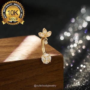 Puede incluir: Un anillo de ombligo de oro con un dije colgante que presenta un grupo de piedras de circonita cúbica transparentes. El dije tiene forma de flor con cinco pétalos. El anillo está hecho de oro de 10 quilates y está adornado con piedras de circonita cúbica transparentes.