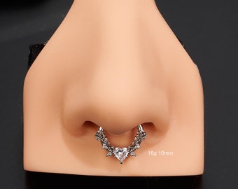 Puede incluir: Un anillo de septum de plata con una gema transparente en forma de corazón y detalles de rosas. El anillo es de calibre 16 y 10 mm de diámetro.  @ouferbodyjewelry