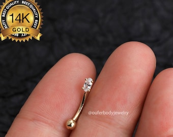 May include: A gold 14K barbell with a clear oval-shaped gemstone. The barbell is held in a hand. The image includes the text "@ouferbodyjewelry".