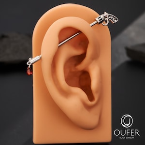 May include: A silver industrial barbell earring with a gun-shaped charm on one end and a red teardrop-shaped charm on the other. The earring is displayed on a tan ear model against a dark background. The jewelry is from OUFER Body Jewelry.