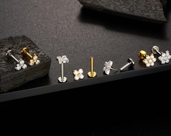 Könnte beinhalten: Eine Auswahl an Lippen- und Ohrpiercings in Blumenform in Silber- und Goldtönen. Einige Stücke sind mit klaren Edelsteinen verziert. Der Schmuck wird auf einer schwarzen Oberfläche vor dunklem Hintergrund präsentiert. Das Bild enthält den Instagram-Handle @ouferbodyjewelry.