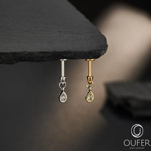 Puede incluir: Dos piercings labret con dijes colgantes en forma de lágrima, uno en metal plateado y otro en dorado, cada uno con una pequeña piedra transparente engastada en la lágrima y el texto OUFER BODY JEWELRY en la esquina.