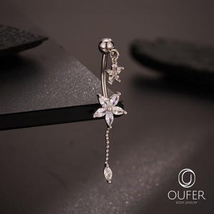May include: A silver-colored belly button ring featuring a floral design. The ring has a curved barbell with a dangling chain and a flower-shaped charm with clear crystals. A smaller star-shaped charm hangs above the flower. The jewelry is set against a dark background.