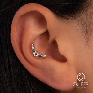 May include: A silver ear piercing featuring a floral design. The jewelry has a curved shape and is adorned with white, green, and red gemstones. The piercing is set against the backdrop of an ear.